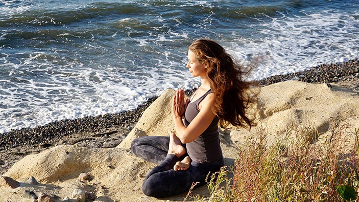 ANJALI MUDRA Frau sitz am Strand, macht Yoga und formt mit den Händen das ANJALI MUDRA