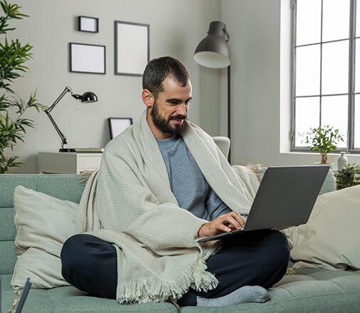 Mann sitzt mit einer Decke um die Schultern geworfen imk Schneidersitz auf einem Sofa und hat einen Laptop auf dem Schoß