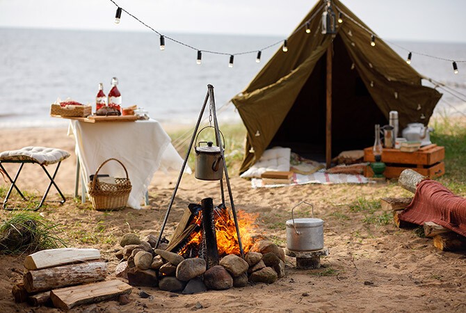Am Strand steht ein Zelt mit Lichterkette, im Vordergrund brennt ein Lagerfeuer und im Hintergrund steht ein gedeckter Tisch