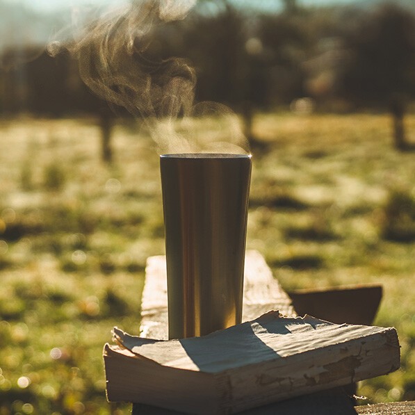 Mehrweg To-Go-Becher Auf einer Bank auf einer Wiese steht ein dampfender Mehrweg-Becher. Davor liegt ein altes Buch.