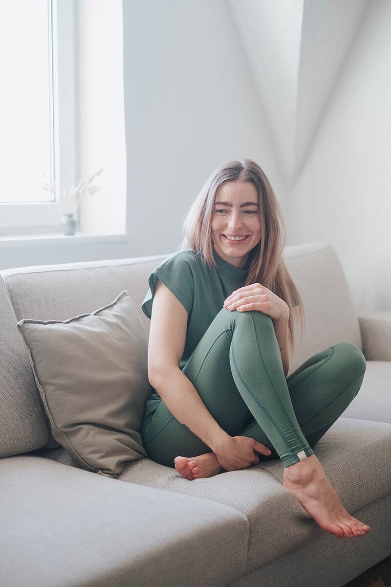 Morgenroutine Ein Mädchen sitzt lächelnd auf einem Sofa. Sie trägt ein chakrana Outfit in Schilf und freut sich über ihre Morgenroutine.