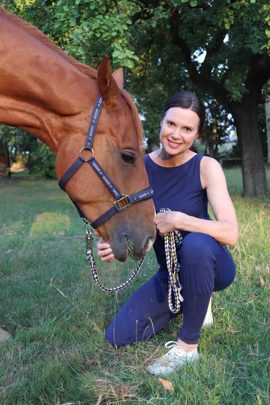 Yoga für Reiter Eine Frau hockt in einem Yoga-Overall auf einem Rasen und hält zärtlich ein Pferd, dass von der Seite ins Bild schaut, am Kopf.