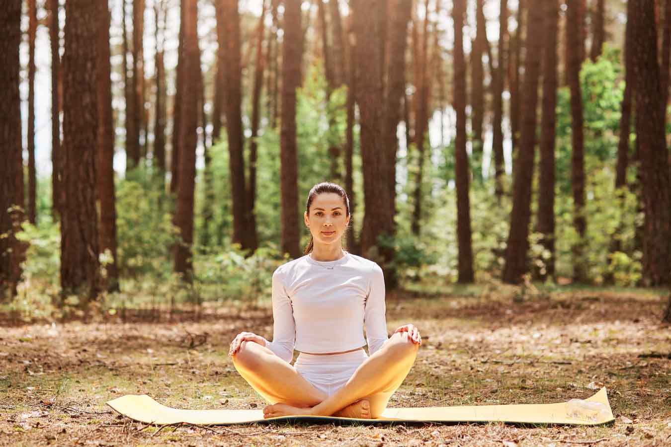 Young beautiful woman with pleasant appearance sitting on karemat in yoga pose and looking directly at camera with calm and relaxed expression, sport training in open air.